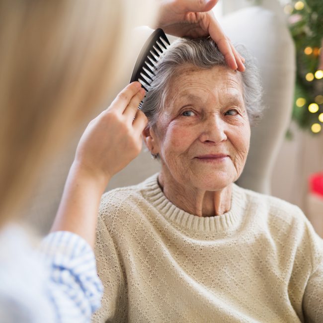 An unrecognizable health visitor combing hair of senior woman sitting on a sofa at home at Christmas time.