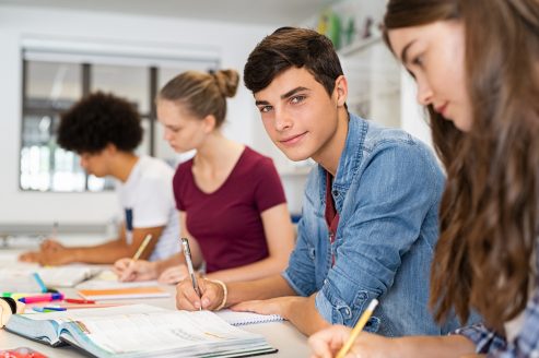 Portrait of happy college young man studying with classmates in library. Group of multiethnic friends studying together while sitting in university classroom. High school guy looking at camera while sitting in a row with girls at desk.