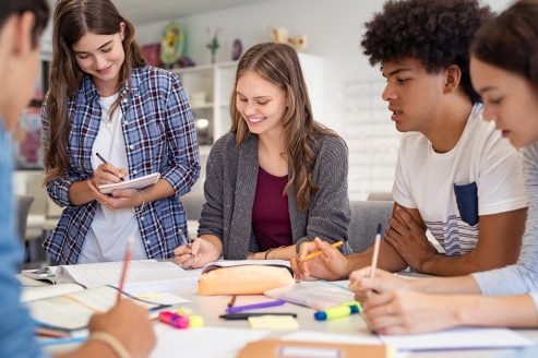 Happy team of high school girls and guys studying together and laughing. Group of multiethnic classmates smiling and studying while sitting in university library. Group of young people sitting at table working on school assignment.