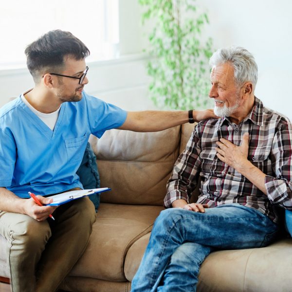 Doctor or nurse caregiver talking with senior man on sofa at home or nursing home